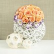 On a crisp white surface, a sophisticated floral centerpiece appears ready to grace a wedding table or elegant dinner party in Tufnell Park. At its heart is a flawless dome of roses, each bloom a tender shade of peach or apricot, opening fully to reveal layered, velvety petals that nestle tightly together. The roses form a near-perfect sphere, a cloud of warm, romantic colour that draws the eye and suggests quiet joy and affection. Around the base of this rose dome, white lisianthus flowers create a soft ruff, their petals edged in vivid purple and deep violet tones that feel almost like watercolour brushstrokes. These frilled edges add drama and refinement, while small sprigs of white baby's breath weave in and out, lending delicacy and a gentle sense of movement, like light wind stirring blossoms in nearby Tufnell Park Gardens. Supporting the blooms is a striking, bulbous container made from white woven rattan or wicker. Its open lattice structure lets light slip through the gaps, casting subtle shadows on the surface below and emphasizing the handcrafted nature of the design. The basket-like vase feels both rustic and contemporary, mirroring Tufnell Park's mix of traditional terraces and creative spaces. In front of the arrangement, two decorative spheres rest on the table: one slightly larger, one smaller, both formed from pale, natural wood-toned strips woven into airy, open frameworks. They echo the texture of the vase and create a pleasing rhythm in the composition. All of this is set against a softly textured beige fabric background, warm and neutral, allowing the peach, white, and violet tones to stand out. The scene suggests an atmosphere of refined celebration and calm beauty, perfect for marking life's quieter milestones and gatherings in North London.