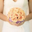 A close-up, chest-level photograph centres on a bride in a classic white wedding gown, her hands gently cupping a perfectly round bouquet of roses and lisianthus, as if paused for a quiet moment just off Tufnell Park Road. The bodice of her dress is trimmed with delicate lace and a soft V-neckline, and the skirt falls in smooth, layered folds that create a clean, luminous backdrop for the flowers. At the heart of the bouquet, countless tightly clustered roses glow in soft peach, warm apricot, and blush pink tones, each velvety petal spiralling inward and catching the light like a late afternoon sky over nearby Hampstead Heath. Below this dome of roses, a striking ring of deep purple blooms with ruffled, white-tipped edges-likely lisianthus-circles the base, forming a vivid, almost painterly contrast that frames the warm centre. Wisps of tiny white filler flowers, reminiscent of baby's breath, peek through here and there, adding a lace-like softness that echoes the gown. Her nails are neatly manicured in a pale, understated shade, with a hint of shimmer on one thumb that mirrors the bouquet's subtle glow. The lighting is gentle and even, with no harsh shadows-just a calm, flattering brightness that suggests a peaceful wedding morning in Tufnell Park, perhaps on the way to a ceremony near the local gardens or a townhouse venue. The overall mood is one of intimate romance and quiet anticipation, capturing the craftsmanship of a careful florist and the tenderness of a bride holding her bouquet close before walking down the aisle.