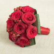 Set against a warm, light beige fabric that resembles delicately woven linen, a compact bouquet of red roses takes centre stage, its perfectly rounded silhouette almost sculptural in its precision. Each rose is in full, luxuriant bloom, petals overlapping in tight spirals that draw the eye inward, then outward again to the gently ruffled edges. The colour palette remains firmly in the family of reds, from bright, ripe-cherry tones to shadowy crimson and deep burgundy that seems to pool in the folds of the petals. Between the blooms, clusters of shiny hypericum berries emerge, their small crimson spheres adding a lively rhythm and a different texture that plays beautifully against the softness of the flowers. Encircling the bouquet, bold dark green leaves have been intricately folded and wrapped, forming a structured green frame that holds the roses as if in a bespoke collar, giving the arrangement a clean, modern finish. The greens have a subtle lustre that catches the overhead light and contrasts gently with the matte background. On the right, the handle of the bouquet stretches out, wrapped in smooth red satin ribbon that matches the roses and is secured so flawlessly it feels almost seamless to the touch. The photograph is taken from slightly above, allowing every detail of the craftsmanship to be clearly seen, the way a bride in Tufnell Park might admire her bouquet before walking through nearby residential streets toward a registry office or a small local church. The atmosphere is one of poised romance, making this design perfect for weddings, milestone anniversaries, or any intimate celebration of enduring love in North London.