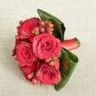 A luxurious, hand-tied bouquet of deep red roses and berries rests on a softly textured light beige fabric, captured from a slightly elevated angle that shows every curve and petal. Around five or six velvety roses sit at the heart of the design, their petals unfurling from tight, shadowed centres into rich layers of crimson and scarlet, like small, glowing lanterns against a neutral backdrop. Threaded between the blooms are clusters of rounded Hypericum berries in shades of reddish-pink and red-orange, each tiny sphere catching the light and adding a playful sparkle to the more formal roses. Broad, dark green tropical leaves are skillfully curved and wrapped around the base, their glossy surfaces and visible veins creating a natural frame that cradles the flowers and adds a sense of movement. At the stems, a smooth red satin ribbon is wrapped tightly and neatly, echoing the rose tones and suggesting the steady hand of an expert florist. The bouquet lies on what appears to be cream or light beige linen, its subtle woven texture softening the scene while making the reds and greens appear even more vibrant. The overall mood is romantic yet composed, suitable as a wedding bridesmaid bouquet, an anniversary surprise, or an intimate gift for someone in Tufnell Park, perhaps after a stroll through nearby streets off Fortess Road or down toward the quiet corners of Tufnell Park Road. The even, gentle lighting emphasises the bouquet's rich colours and layered textures without harsh shadows, giving the impression of fresh, cool petals and foliage arranged just moments ago, ready for local delivery across this North London neighbourhood.