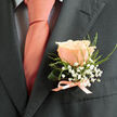 This boutonniere is a study in refined colour and thoughtful construction, crafted to sit perfectly on a charcoal lapel. A flawless peach rose commands the centre-its petals soft and layered, carrying warm coral-pink tones that reflect the sheen of the silk ribbon securing the stems. Surrounding the bloom, delicate clouds of white baby's breath create an ethereal frame, while fine green foliage and a curled supporting leaf bring a liveliness and subtle architectural curve to the piece. The rose and a pale pink tie read as a matched pair, their warmth mellowed by the suit's deep tone so every detail is readable and harmonious.

The tactile contrasts are easy to imagine: the rose's velvet surface, the airy texture of gypsophila, the cool smoothness of satin ribbon and the crisp structure of fresh greenery. A faint, pleasant scent seems implied, the kind that lifts the spirit at a wedding or anniversary. As a local florist mindful of the community around Tufnell Park and Fortess Road, I often assemble buttonholes like this for couples walking from the tube toward small family venues in the area. Flowers Tufnell Park prepares each piece with secure wiring and a neat finish so it endures a day of embraces and photographs, offering both a visual flourish and a tender, wearable memory.