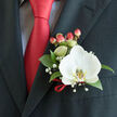 A single, luminous Phalaenopsis orchid anchors this boutonnière with quiet authority, its white petals broad and satiny and a yellow-gold patterned labellum at the centre that reads like a tiny work of art. The flower is mounted against a dark charcoal jacket lapel, the contrast sharpened by a solid red satin tie whose smooth sheen is echoed in a slender ribbon tied around the base of the stems. Deep green, glossy leaves cradle the bloom, their firm texture giving structure to the arrangement, while delicate sprigs of gypsophila weave through like soft breath - tiny stars that add nostalgic lightness. A small cluster of hypericum berries, coloured a coral-tinged red, provides a rounded counterpoint and picks up the tie's hue so the eye travels seamlessly from personal style to floral detail. Beneath the lapel, a subtle light-blue or grey grid shirt peeks through, suggesting the thoughtful coordination of attire often chosen for weddings, proms, or formal galas. The orchid's cool silkiness and the ribbon's satiny warmth are captured under gentle, diffused light that emphasises texture without glare. Created with the same attentive hand that local couples trust in Tufnell Park, this boutonnière would sit comfortably on a groom leaving the tube station on Tufnell Park Road or on a guest arriving from nearby Fortess Road - a refined, wearable accent that carries the freshness of a florist's care and the quiet confidence of well-chosen details. Flowers Tufnell Park can recreate this look for local deliveries when time and season allow.