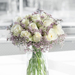 A softly lit bouquet of white roses and lavender statice stands in a clear cylindrical glass vase, centred on a clean white surface that makes the blooms appear almost luminous. The roses, perhaps two dozen in number, are gathered into a full, rounded composition; each blossom is a creamy white, with petals curling inward in neat spirals, the outer layers just touched by a hint of pale green that betrays their freshness. Interlaced with these roses are delicate sprays of statice in varying shades of lavender and muted purple, their tiny star-like flowers adding a veil of texture that wraps gently around the more solid rose heads. The vase is simple yet elegant, transparent enough to reveal the straight green stems arranged with care and submerged in clear, light-catching water. Soft natural light, reminiscent of a quiet morning along Tufnell Park's tree-lined streets or a calm afternoon near the local station, drifts across the scene, leaving subtle highlights on the petals and casting faint shadows that suggest depth rather than drama. Behind the arrangement, the background dissolves into an abstract wash of light grey and white, hinting at an airy, uncluttered room where the bouquet is given space to breathe. The overall sensation is one of stillness and solace, the cool palette of whites and purples creating a feeling of dignity and composure. This is the sort of arrangement you might find resting on a sideboard during a memorial gathering, or delivered to a loved one near Tufnell Park as a quiet expression of sympathy, offering comfort through its simple, steadfast beauty.