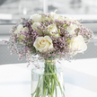 A graceful sympathy bouquet is captured in this close-up image, its calm beauty echoing the quieter streets around Tufnell Park. At its centre, numerous white roses in full bloom unfold velvety petals in soft spirals, with tones that drift from pure white to warm cream and faint ivory. These roses feel both dignified and tender, their generous blossoms suggesting a gentle embrace. Surrounding and weaving between them is a haze of tiny lavender and pale purple filler flowers, most likely statice, forming a soft, cloud-like veil. Their many small, papery florets introduce a subtle variation in texture, giving the arrangement an ethereal, comforting quality-something you might send when words are hard to find. All of the stems are gathered neatly into a tall, clear glass vase filled with fresh water, allowing the long green stems to remain visible as they criss-cross in quiet, natural lines. The vase rests on a smooth white surface that reflects some of the ambient light, reinforcing the bouquet's luminous character. In the background, everything falls gently out of focus into pale grey and white tones, suggesting a simple, modern interior, perhaps a peaceful front room near Fortess Road or a light-filled hallway on a side street off Brecknock Road. Soft daylight glides across the roses and filler flowers, creating a tranquil glow without sharp contrasts. Overall, the arrangement radiates a feeling of reverence, calm, and sincere condolence, ideal for offering comfort and remembrance to families across Tufnell Park, Archway, and the nearby North London community.