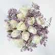 This photograph shows a tranquil, circular bouquet captured from directly above, resting against a pure white background that highlights every petal and stem. Dominating the arrangement are flawless white roses, around a dozen in number, their petals cool and velvety to the eye. Some of the roses are tight and reserved, holding their shape like small, perfect cups; others have begun to open, revealing soft spirals that deepen to a faint ivory glow at their centre; a few are fully open, with loose, layered petals that create intricate patterns and subtle shadows. Encircling and weaving through these roses is a cloud-like ring of lavender limonium, the tiny clustered flowers forming a gauzy, almost shimmering border in muted purple. This limonium, sometimes called sea lavender, brings a gently textured contrast that feels both airy and embracing, as though the roses are held within a protective haze. Occasional glimpses of green stems and delicate foliage appear beneath the blooms, contributing a quiet, natural freshness without breaking the soothing white-and-lilac harmony. The overall effect is one of gentle stillness, reminiscent of a peaceful moment in a small Tufnell Park garden after rain, when colours appear soft and subdued. It is easy to imagine this bouquet being delivered to a home near Tufnell Park Underground as a heartfelt condolence gift, placed on a table beside a framed photograph. With its careful balance of purity, softness, and subtle colour, the arrangement feels designed to offer comfort during times of loss, supporting loved ones through sympathy, remembrance, and the reassurance of simple, enduring beauty.