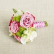 A small, exquisitely arranged hand-tied bouquet lies horizontally on a light tan, textured fabric that looks like natural linen spread over a table in a Tufnell Park townhouse. Two prominent, fully opened pink roses form the heart of the design, their soft blush petals deepening to a more intense magenta along the ruffled outer edges, creating a gentle ombre effect as the spiralled petals curl towards tightly furled centres. Nestled between and around them are compact tufts of bright, apple-green button chrysanthemums, each tiny floret forming a plush, spherical pom-pom that adds playful texture and a modern splash of vivid colour. Creamy white filler flowers, possibly petite lisianthus or waxflower, weave in and out of the composition, their dainty, multi-petalled blooms catching the soft, even light and giving the bouquet an airy, romantic lightness. Discreet hints of darker green foliage peek from beneath the blossoms, adding depth and a natural garden feel, like something freshly gathered from a North London allotment. The neatly trimmed stems are bound together in a smooth, iridescent fuchsia-pink satin ribbon, which forms a polished handle that would sit comfortably in the palm of a bridesmaid walking through nearby Hampstead Heath pergola gardens for photos. A delicate strand of tiny white faux pearls spirals diagonally over the ribbon, as though scattered dewdrops have settled there, lending a subtle vintage glamour. Resting against the neutral, lightly woven background, the bouquet's pink, green, and white palette appears vivid yet refined, perfect as a bridesmaid's posy, an intimate bridal bouquet, or a charming centrepiece for a small wedding breakfast in a Tufnell Park café, capturing both classic elegance and fresh, contemporary London style.