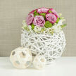 On a clean, reflective white tabletop, a thoughtfully styled floral centerpiece captures the gentle romance of a modern Tufnell Park wedding breakfast. The eye is first drawn to the unusual vase: a hollow sphere woven from slender branches painted soft white, like a sculptural bird's nest reimagined for a contemporary Camden townhouse. Through this airy lattice, you glimpse stems and foliage supporting an abundant dome of pastel flowers. At the crown, rounded pink roses unfurl in full bloom, some petals shaded with slightly deeper, almost blush-mauve tones at the edges, giving depth and softness. Nestled confidently among them are plump lime-green button chrysanthemums, their small, rounded heads offering a crisp contrast of colour and texture, as if tiny mossy cushions have been tucked between the roses. Encircling the lower edge, clusters of tiny white blossoms with faint yellow centres spill lightly over the woven rim, blending the structured sphere with the organic life it holds. Beside the arrangement, resting on the same white surface, sit two decorative balls crafted from pale wooden strips twisted into loose lattices, the larger one echoing the central vase's form and the smaller adding a playful rhythm. Behind everything lies a gently blurred wall in warm beige tones, reminiscent of woven fabric, which enhances the softness of the scene. The composition suggests natural daylight filtering through, highlighting the petals' velvety texture and the twig vase's papery finish. This design feels serene and balanced, perfect for couples marrying close to Tufnell Park Station or hosting a refined celebration at home, where flowers are chosen to express tenderness rather than excess.