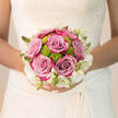 Held close against the intricate lace of a bridal gown, this round, hand-tied bouquet captures the intimate, joy-filled mood of a wedding morning in Tufnell Park. At the heart of the design, lush roses in a soft blend of lilac and dusky pink are fully open yet still resilient, each bloom formed from dozens of gently curling petals. Between them sit plump clusters of lime-green button chrysanthemums, their tidy, spherical heads giving a playful, contemporary twist to the otherwise timeless arrangement. Along the outer edge, creamy white flowers and pointed ivory buds trace a soft border, like a band of floral lace echoing the bride's dress as she stands near the leafy streets around Tufnell Park station. The colour palette is calm but uplifting: muted mauve and blush tones balanced by fresh green and clean white, creating an overall effect that feels like early daylight filtering through a garden. You can almost imagine the scent: the familiar sweetness of rose layered with the cool, green aroma of newly cut stems, a fragrance that steadies the breath and heightens anticipation. The florist's craftsmanship shows in the neat spiralling of stems, the careful spacing of each bloom, and the way the bouquet sits naturally in the bride's hands, ready for photos in nearby Waterlow Park or a ceremony in a local church. Designed to last from the exchange of rings to final farewells, this bouquet becomes a tangible memory of joy, hope, and new beginnings, expressed in petals, foliage, and light.