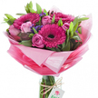 A radiant hand-tied bouquet sits upright in a clear cylindrical glass vase, the bundled stems visible through the water and bound with a simple rustic twine knot that holds a small white rectangular tag stamped with a stylised pink-and-green rose logo. At the heart of the arrangement, several large gerbera daisies flare in vivid fuchsia with dark, almost black centres that anchor the brightness and draw the eye inward. Surrounding them are soft pink roses in a gentle orchestra of stages-tight, elegant buds promise future bloom while fully opened roses display silky, layered petals in a pastel blush. Small clusters of deep purple-blue filler flowers, likely statice, thread through the composition, adding a fine, textured counterpoint to the broader petals and lifting the colour palette toward jewel tones. Lush greenery-broad smooth leaves and slender stems-forms a verdant backdrop, giving the bouquet a natural depth and a fresh, garden-picked feel. The blooms are cradled in crinkled, sheer pink wrap that graduates from a near-translucent pale pink halo around the blossoms to a more saturated, celebratory pink lower down, catching the light with a soft sheen. The overall mood is uplifting and intimate, a creation that speaks of thoughtful giving and seasonal cheer. As a local florist I imagine this piece brightening a table in a Tufnell Park flat or arriving as a birthday surprise near Fortess Road; it smells faintly sweet and floral, visually sings in pinks and purples, and reads as a joyful, hand-crafted gift for someone you want to make smile.