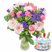 A bright, celebratory bouquet sits in a clear glass vase, captured against a crisp white background with a lively birthday accent to the lower right. The hand-tied arrangement opens with several elegant pink roses-petals layered like silk in shades from soft blush to warmer pink-each bloom full and romantic. In front and just below, pale pink Gerbera daisies fan their broad, ray-like petals around butter-coloured centres, giving the bouquet a playful, friendly energy. Scattered through the flowers are clusters of vivid purple filler blooms, likely statice or limonium, their tiny, papery florets adding a fine, textured contrast that deepens the composition. Small white spray blooms-suggestive of spray carnations or alstroemeria-nestle into the mix, lending moments of brightness and fullness between the larger focal flowers. Glossy deep-green foliage frames and supports the blossoms, the leaves varying in shape and sheen so the whole arrangement reads as lush and natural. Through the glass vase the green stems and clear water are visible, a simple detail that underscores freshness. To the lower right a small foil cake-shaped birthday balloon adds a cheerful note: a white frosted top with colourful sprinkles, a pale blue base, and the words 'Happy Birthday' printed in multicoloured letters. Four tiny candles in red, yellow, green and blue, each with a small flame motif, perch on the cake, reinforcing the festive mood. The overall impression is one of thoughtful celebration-an arrangement that smells faintly sweet, looks freshly gathered, and would be an easy choice for a local birthday surprise delivered within the Tufnell Park neighbourhood or handed over at the nearby tube or Fortess Road market.