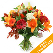 A hand-tied autumn bouquet captured against a clean white studio background, the composition glowing with warm seasonal colour and confident florist craft. At the heart of the arrangement are wide-open trumpet-shaped orange lilies, their smooth petals and pollen-dusted stamens providing a dramatic, aromatic focal point; a few slender lily buds push upward, promising a second wave of blooms. Ringing the lilies are plump Gerbera daisies in deep red and bright orange, their layered disks of petals offering cheerful, sculptural faces. Soft white Lisianthus with gently ruffled edges thread through the composition, their creamy tones calming the palette and lending a delicate, tactile contrast to the bolder flowers. Small, star-like Astrantia with purple-pink centres scatter among the stems, giving fine, lacy texture that invites a closer look. Dark, glossy foliage-likely ruscus or salal-builds a lush green foundation and frames the blooms, while the most striking seasonal detail is the inclusion of large, deeply lobed oak leaves in shades of burnt orange and reddish-brown, placed like confetti to reinforce the autumnal mood. The stems are neatly gathered and bound with dark twine or raffia in a traditional hand-tied style, suggesting freshness and ease of display. Soft, diffuse lighting highlights the velvet of each petal and the crisp edges of the oak leaves, evoking a cool October morning. In the lower right corner a bright orange diagonal banner reads FREE CHOCOLATE in bold white letters, a visible promotional accent. Prepared by experienced local florists for deliveries across Tufnell Park and nearby Kentish Town, this bouquet suits Thanksgiving greetings, birthdays, or simply a seasonal, fragrant surprise for a doorstep.