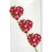 This image shows an elegant trio of funeral hearts, each one carefully composed from hundreds of red and pink roses, cascading diagonally down a pristine white surface as though arranged along a tranquil chapel wall in Tufnell Park. The hearts are stacked vertically, with the top heart slightly to the left and the lowest heart somewhat to the right, creating a gentle, flowing line that draws the eye downwards. Every heart is densely packed with roses that range from intense crimson to vivid fuchsia and softer blush pinks, their velvety petals overlapping in layers to form a plush, almost cloud-like texture. Some blooms are fully open, revealing intricate spirals at their centres, while others remain partially closed, adding subtle shadows and dimension. Encircling and linking the hearts, slender ivy vines weave a continuous thread of deep green, their small, glossy leaves outlining the shapes and trailing gracefully between them. These natural green lines create a sense of unity, as if the hearts are joined by invisible ties of memory and affection-much like families and friends connected across Tufnell Park, Archway, and nearby streets. The background is bright, smooth, and white, reflecting cool, even light that makes the colours appear clean and vivid without distraction. A faint vertical pattern and a soft side shadow hint at a carefully prepared display panel, reminiscent of a modern funeral venue or remembrance space. The overall mood is tender and reverent, a romantic symbol of enduring love transformed into a tribute suitable for funerals, memorials, or celebrations of life where every rose can be read as a silent message of sorrow, gratitude, and unbroken bond.