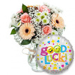 A cheerful, carefully composed 'Good Luck' gift set photographed against a clean white background so every pastel tone and shiny surface reads clearly. On the left a generous bouquet sits in a simple clear glass vase, the neck finished with a rustic twine bow; the stems are visible through the glass and the arrangement feels both hand-tied and generously styled. Soft peachy-orange Gerbera daisies with distinctive dark brown centers mingle with light pink, tightly furled roses whose petals suggest a tender, rosy perfume. Dainty sprays of white baby's breath (Gypsophila) thread through the bouquet like lace, while clusters of small white and lime-green chrysanthemum florets add crisp texture and a lively, green-tinged sparkle. Deep green foliage peeks between the blooms, giving the composition depth and a natural rhythm. To the right and overlapping the lower portion of the vase, a round metallic silver foil balloon catches the light: its reflective surface is patterned with tiny multicoloured stars, hearts and confetti shapes in blue, pink, yellow and orange. The playful message 'Good Luck!' stretches across the balloon in bouncy, multi-coloured letters - the two o's in 'Good' replaced by yellow smiley faces, one winking and the other sticking its tongue out - and a small KALEIDOSCOPE logo sits unobtrusively at the balloon's base. The overall impression is encouraging and celebratory: a balanced pairing of fresh, soft-hued flowers and a buoyant, optimistic balloon that reads as a thoughtful send-off or a comforting boost for exams, interviews or new beginnings around Tufnell Park and nearby Dartmouth Park.