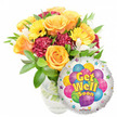 A warm, uplifting get-well ensemble seen against a clean white backdrop: to the left, a lush glass vase brims with a generous hand-arranged bouquet that feels like a small sunlit garden. Several large, soft orange-to-deep-yellow roses open in layered spirals, their velvety petals catching the light and offering a faint citrus-sweet scent that lifts the spirit. Bright yellow gerbera faces push cheerfully through the composition, their flat, sunny discs contrasting with the ruffled texture of deep burgundy or maroon carnations tucked between stems. Delicate white alstroemeria peek through in clusters, their fine markings and fragile petals adding an airy, graceful punctuation among the bolder blooms. Abundant glossy green foliage-long willow-like leaves and rounded greenery-cradles each blossom, giving the arrangement a fresh, garden-picked silhouette. The stems and clear water are visible through the vase, lending a simple, honest freshness. Nestled slightly overlapping at the lower right, a shiny silver Mylar balloon plays a joyful counterpoint: its reflective surface frames a playful cluster of cartoon-style balloons in blue, purple, green, pink and yellow, some patterned with stripes and polka dots, while bold yellow-and-blue lettering proclaims Get Well Soon. Small starburst graphics and a tiny KALEIDOSCOPE logo at the balloon's edge complete the cheerful message. Bathed in soft daylight-similar to the gentle morning light on Tufnell Park Road-this arrangement conveys comfort, optimism and the tactile reassurance of a thoughtful visit from a neighbour or friend during recovery.