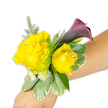 A hand-crafted wrist corsage rests diagonally across a light-to-medium skinned forearm against a crisp white background, its composition designed to read clearly from left to right. Two buttery yellow roses form the cheerful heart of the piece: one open and expansive to the left, the other slightly cupped and intimate to the right, each petal layered with the soft, satiny texture of garden roses. Small, clear rhinestone studs nestle near the centre of both blooms, catching studio light and adding a refined, celebratory glint with every subtle movement. Behind and rising above the roses a single, sculptural calla lily unfurls in a deep plum-burgundy so dark it reads almost black at the throat, offering a dramatic, velvety contrast that gives the corsage evening polish. Fine asparagus-fern threads and feathery green sprigs weave through the composition for air and movement, while rounded variegated leaves-pale at the margins, richer green at the centre-form a steady base and cool the palette. Clusters of tiny white filler flowers, reminiscent of spray chrysanthemums or miniature gypsophila, are tucked into hollows to create fullness without weight. The wristband is mostly concealed beneath the blooms, the arrangement secured so it sits comfortably and diagonally from the lower left of the photo toward the middle right, showing how the piece will read on the arm. The whole effect is a blend of sunny warmth and sophisticated dusk tones, scented softly with rose and green foliage; a wearable accent suited to prom, bridal party duties, or a guest marking a special evening in Tufnell Park or the neighbouring Dartmouth Park.