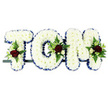 This image features a personalised floral name tribute reading TOM, a three-letter sympathy arrangement designed with care that would look fitting at a funeral or memorial in the Tufnell Park area. Each letter is shaped in bold block capitals, arranged side by side on a slim support bar that is almost hidden beneath the flowers, set against a bright white background that emphasises every detail. The surfaces of the T, O, and M are densely packed with pristine white chrysanthemums, their uniform, button-like blooms forming a thick, cushiony layer that feels soft to the eye and symbolises purity and remembrance. Encircling each letter is a royal blue satin ribbon border, carefully ruffled to create gentle folds and give a crisp outline that contrasts gracefully with the snowy petals. At carefully chosen points on the letters, you can see small clusters of accent flowers: rich red roses, some fully open and others in tight buds, delicate white and cream spray roses, and slender white freesia-style stems emerging from tufts of lush greenery. Fern fronds and glossy leaves add movement and a natural, garden-like feel, softening the structured shape of the letters. The red, white, and blue palette carries quiet symbolism-love, peace, and respect-while the overall design speaks of a bespoke farewell made in honour of Tom. It is easy to imagine this tribute prepared by Flowers Tufnell Park and delivered with care to a local crematorium, church, or family home near Junction Road, offering a dignified final message in flowers.