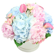 A lavish pastel bouquet spills generously from a round white hatbox, photographed against a bright white background that lets every colour and texture stand out. The arrangement is dense and full, dominated by large, romantic pink peonies whose layers of soft, ruffled petals create a sense of luxurious depth. Around and among them, pale blue hydrangeas form cushiony clusters, each one made up of countless little blossoms that give the design a pillowy, cloud-like feel and introduce a cool, calming tone. Small light pink spray roses thread through the larger flowers, some still tightly closed and others beginning to unfurl, adding fine detail and a sense of unfolding time. Rising gracefully from the pastel mass are two striking calla lilies in a more intense shade of pink, one positioned towards the upper left and the other on the right, their smooth, trumpet-shaped blooms introducing a sleek, contemporary contour. A few dark green leaves are just visible near the base, peeking over the rim and hinting at fresh stems nestled inside the container. The hatbox itself is simple and elegant, round and spotless with a faint ridge echoing its circular form, the kind of stylish presentation you might see delivered to a terrace house near Tufnell Park station or to a studio along Fortess Road. Soft, even lighting washes over the scene, making the pastel blues and pinks appear especially gentle and soothing. The overall impression is of refined joy and thoughtful beauty, ideal for celebrating a new arrival in N19, sending get-well wishes, or providing a quietly sophisticated centrepiece for a family gathering in Tufnell Park.