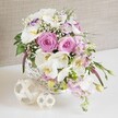 An opulent yet softly romantic floral centerpiece, perfect for a wedding reception table in Tufnell Park, is arranged in a low, round white wicker basket placed on a smooth white surface. The composition overflows with pastel pink and pure white flowers, creating a gentle cascade of colour and texture. At the forefront, fully opened pink roses reveal tightly layered spiral petals, their velvety surfaces suggesting a subtle fragrance and a sense of timeless romance. Around them, elegant white Phalaenopsis orchids fan out, their rounded petals gleaming almost like porcelain, each flower marked by a warm yellow-orange centre that draws the eye inward. Ruffled white Lisianthus with delicate lavender-tinted edges lend an extra layer of sophistication, while stems of soft pink stock rise and drift slightly over the edge of the basket, adding movement and height. Tiny clusters of white baby's breath are scattered between the larger blooms, creating a misty, ethereal effect that softens the transitions and enhances the bridal feel of the arrangement. Glossy green leaves and a few gracefully trailing buds introduce fresh, natural contrast, hinting that the flowers have been carefully selected at peak freshness. In front of the basket, two decorative balls made of pale, intertwined wicker or rattan rest together, their open latticework harmonising beautifully with the basket's weave. Behind the arrangement, a neutral, linen-like backdrop in light beige or taupe keeps the focus firmly on the flowers, evoking the calm, elegant atmosphere of a small wedding or anniversary dinner in one of Tufnell Park's cosy venues. The overall impression is one of gentle luxury, handcrafted detail, and celebratory warmth.