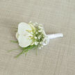 A refined white orchid boutonnière is presented on a pale, linen-like fabric, its calm beauty captured in soft, natural light that could easily be streaming through a townhouse window in Tufnell Park. At the heart of the design sits a single orchid bloom, perfectly formed, with broad, velvety petals that curve gently outward. Their pure white surface is almost luminous against the neutral background, while the centre of the orchid reveals a delicate touch of buttery cream and soft yellow, suggesting warmth and intimacy. Surrounding the orchid, downy sprays of white baby's breath cluster together, each tiny flower creating a misty, romantic halo, adding volume without overwhelming the central bloom. Fine strands of slender green foliage emerge from beneath, their bright, fresh tone reminiscent of park grass after rain in nearby Dartmouth Park, bringing a sense of life and movement as they fan lightly to one side. The stems are meticulously gathered and wrapped in a band of smooth white satin ribbon, its subtle sheen catching the light in quiet highlights. The ribbon is finished with a compact, symmetrical bow, tied high on the handle so the wearer's fingers would meet only soft satin when pinning it in place. Beneath the boutonnière, the woven beige fabric shows a faint, regular texture, offering a gentle contrast that keeps the arrangement from appearing flat, almost like a carefully chosen swatch of suit cloth from a local tailor. The atmosphere of the image is poised and tranquil, suggesting a wedding morning just beginning - jackets hanging ready, ties being straightened, and this orchid boutonnière waiting to become the final, elegant touch for a groom or best man heading out to a ceremony close to Tufnell Park Underground.