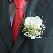 In this image, a single pocket boutonnière becomes the poetic focal point of a sharply tailored suit. A white orchid, flawless and serene, unfolds at the centre, its petals gently cupped as though holding a secret. Their texture appears silky and cool to the touch, glowing softly against the dark weave of a charcoal jacket. At the heart of the orchid, a hint of buttery gold adds depth, suggesting warmth and intimacy. Surrounding the bloom, clusters of baby's breath form a delicate halo, each tiny floret like a suspended pearl of light. Wisps of green foliage add freshness and a sense of movement, echoing the leafy streets around Tufnell Park and the natural calm of nearby green spaces. At the base, a neat white ribbon has been tied with careful precision, its satiny surface catching the light and hinting at the meticulous craftsmanship of a local Tufnell Park florist. The boutonnière rests just above the pocket, framed by crisp stitching and the bold slash of a red tie, whose rich colour adds drama and contrast to the composition. Against this backdrop, the flowers feel almost luminous, celebrating tradition without feeling overly formal. The mood is quietly romantic, evoking a wedding day stroll through the neighbourhood, perhaps en route to a ceremony near Archway or Gospel Oak. You can almost sense the anticipation in the air, the rustle of suit fabric, the faint fragrance of fresh blooms, and the way a small, considered floral piece can hold so much meaning on a day dedicated to promises and shared futures.