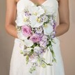 Framed in the gentle light reminiscent of a spring afternoon near Tufnell Park's leafy streets, the bride's arms curve protectively around a spectacular cascading bouquet. White Phalaenopsis orchids take centre stage, gleaming softly with their wide petals and subtle yellow throats, channeling the refined serenity often found at intimate Tufnell Park wedding venues. Surrounding the orchids, blushing garden roses open in full, their pale pink and mauve tones evoking both modern romance and vintage charm. Delicate lavender stocks and clusters of lisianthus-some with ruffled edges tipped in a dusky purple-thread through the arrangement, infusing movement and texture, much like floral displays seen along Brecknock Road. Wisps of baby's breath wash the bouquet in airy white, while occasional strokes of greenery provide depth without overwhelming the gentle palette. The bridal gown, strapless and intricately laced, provides an elegant frame for the bouquet's volume and cascading form. Every bloom appears thoughtfully placed, their freshness reflecting the expertise of Tufnell Park's local florists and the aesthetic sensibilities cherished throughout North London. The image as a whole conveys an atmosphere of romantic anticipation-timeless and graceful, designed for couples seeking exceptional wedding flowers and lifelong memories.