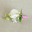 A delicate white orchid boutonniere is laid horizontally on a softly textured, light beige linen-style cloth, the neutral background allowing every detail of the design to stand out with calm clarity. At the centre, a single Phalaenopsis bloom opens wide, its broad, velvety petals a pure, luminous white that seems to catch the gentle, even light drifting across the scene. In the heart of the orchid, a small flare of soft yellow and the faintest touch of pale pink create a subtle warmth, like a whisper of colour against ivory silk. Clusters of tiny white baby's breath are tucked closely around the base, their cloud-like sprays adding a feathery lightness and a sense of romantic haze, reminiscent of spring weddings in Tufnell Park's quiet streets. Slender, fresh green leaves peep from behind the flowers, introducing a cool, natural contrast that keeps the piece feeling lively and freshly made. To one side, a short stem carries tightly closed, lime-green orchid buds, their smooth surfaces hinting at flowers still to come, a quiet symbol of new beginnings and promises. The stems are neatly bound together with a slim, bright fuchsia pink satin ribbon, tied into a precise little bow whose glossy sheen brings a joyful pop of colour, ideal for a groom or prom guest stepping out near Tufnell Park station or along Fortess Road. The overall effect is one of refined simplicity and timeless elegance, a floral accessory that feels intimate, personal, and thoughtfully crafted for a special day.