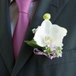 A close-up photograph reveals a carefully tailored charcoal suit jacket in soft, even light, its left lapel framing an elegant white orchid boutonniere that seems to glow against the dark fabric. The jacket is cut from a finely woven cloth, the texture smooth and refined, with a crisp edge that leads the eye down toward a neatly folded breast pocket. Just beside it, a mauve or deep plum tie lies perfectly knotted over a pale grey dress shirt, whose subtle pattern adds quiet interest without distracting from the flowers. Pinned to the lapel is a single Phalaenopsis orchid bloom, its broad, satiny white petals gently curving outward to expose a warm centre brushed with yellow and a hint of soft orange. Above this open flower sits a closed, pale green orchid bud, suggesting new life and the promise of the day ahead. Around the orchid, delicate sprigs of lilac-toned blossoms, reminiscent of heather or another fine-textured flower, mingle with airy clusters of white gypsophila, known as baby's breath, creating a cloud-like halo. Slender, deep green leaves give the piece structure and contrast, their smooth surfaces catching the light. At the base, a slim lavender satin ribbon is tied into a neat bow, its sheen echoing the silk of the tie and suggesting thoughtful coordination. The composition feels calm, polished, and romantic, like a groom standing ready at a Tufnell Park ceremony, perhaps just moments away from walking into a local church or registry office near the park. The image captures that quiet pause before celebration, when flowers, fabric, and emotion all come together in one understated yet unforgettable detail.