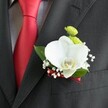 A close-up photograph reveals a beautifully crafted wedding boutonniere pinned neatly to the lapel of a dark charcoal grey suit jacket, the fine wool fabric showing a subtle, woven texture that catches the soft indoor light. Just to the left, a vivid red satin tie runs down the centre of a light grey and white small-checked dress shirt, its smooth, almost mirror-like sheen adding a bold streak of colour that hints at celebration. Nestled against the lapel, the boutonniere is built around a single, large white Phalaenopsis orchid, its broad petals velvety and serene, cupping a delicate yellow-and-white lip at the centre. This orchid sits like a tiny sculpture, the focal point of the arrangement, its pure white tones glowing gently against the dark suit. Surrounding it, airy clusters of tiny white baby's breath create a halo of softness, each miniature bloom adding a sense of romance and lightness, reminiscent of fine mist drifting over nearby Tufnell Park Fields on an early spring morning. Two small, rounded lime-green chrysanthemum-style button blooms peek out just above the orchid, their bright, fresh green adding playful contrast and a touch of modernity. A few broad, glossy green leaves frame the flowers, adding depth and a natural, garden-fresh feeling, as though the stems had just been prepared in a local studio off Fortess Road. At the base, a narrow deep red satin ribbon wraps the stems precisely, its hue perfectly echoing the groom's tie and drawing the whole composition together. The boutonniere sits just above a neatly stitched jacket pocket, a small yet meaningful floral accent that enhances the formality of a wedding or special celebration in Tufnell Park, radiating quiet confidence, love, and careful craftsmanship.
