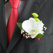 This image captures a refined wedding boutonnière pinned to a classic dark suit, the kind of thoughtful finishing touch you might see at a ceremony near Tufnell Park's leafy streets. At its heart rests a pristine white orchid, its petals satiny and thick, unfurling in a gentle curve that feels both sculptural and serene. A soft hint of buttery yellow glows from the orchid's centre, drawing the eye in and adding warmth to the otherwise cool, white surface. Behind this focal bloom, petite lime-green button chrysanthemums cluster together, their rounded, tightly packed petals creating a lively texture that suggests new beginnings, much like fresh foliage in nearby parks. Wisps of baby's breath weave delicately among the flowers, each tiny white blossom like a dot of lace, spacing out the composition and giving it a weightless, cloud-like frame. Dark, glossy leaves provide a grounding contrast, their deep green tones quietly supporting the brighter blooms above. At the base, a slim band of deep red ribbon is wrapped with care, its rich colour mirroring the silk tie on the wearer and adding a romantic accent that feels perfectly suited to a Twickenham or Tufnell Park wedding. The lighting appears soft and even, highlighting the natural sheen of each petal and leaf without harsh shadows. Everything about this boutonnière speaks of subtle craftsmanship and intention, from the clean lines to the harmonious colour palette, creating an elegant floral accent that will stay close to the wearer throughout their vows, photographs, and evening toasts.