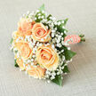 There is a hushed, almost springlike magic captured in this bridal bouquet: six creamy peach roses cluster at its heart, their petals spiralling outward with a delicate luminosity that invites a gentle touch. Nestled around them, clouds of white gypsophila create a frothy, fairy-like border that lifts and lightens the composition, making the roses appear to float within the bouquet. Sprays of glossy emerald foliage thread through the flowers, offering structure and a cool visual counterpoint to the warm peach tones. The handle is swathed in matching peach satin ribbon, its smooth surface catching soft light, and finished with a slender string of pearls that adds an elegant, heirloom rhythm to the piece.
Held, the bouquet feels reassuringly light and fragrant, with the roses' velvet petals releasing a tender, green-tinged scent that suggests garden mornings rather than heavy perfume. I imagine a bride carrying this down a leafy street in Tufnell Park, past the cafés that line Junction Road or beneath the dappled shade of Dartmouth Park's trees, the bouquet offering a quiet glow against her dress. Its craftsmanship is precise yet gentle - a design that works as ceremony décor, a bridal keepsake, or an intimate gift that conveys devotion and hope. The overall impression is of refined simplicity: a timeless arrangement made to celebrate beginnings with graceful understatement.