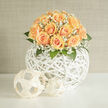 Peach-toned roses form a perfect, rounded bouquet resting on a sculptural white wicker orb, each bloom layered with tender, velvety petals that graduate from creamy peach to faint apricot at the heart. Sprays of delicate white gypsophila braid through the roses like soft confetti, their tiny blossoms scattering light and creating a dreamy, veil-like quality that lifts the composition. Fresh green foliage frames the dome, the leaves' cool tones setting off the warmth of the roses and adding a subtle architectural line to the overall shape. The vase is more sculpture than vessel: a spherical, open-weave form crafted from white rattan that gives the bouquet a modern, airy silhouette and allows gentle sunbeams to filter through and play across the blooms. Imagine this arrangement catching late afternoon light on a café table near Tufnell Park Road or bringing a serene presence to a mantel on Fortess Road; it feels at home in both contemporary interiors and intimate celebration settings. The scent is quiet and tender-soft rose with a hint of green freshness-perfect for weddings, newborn visits, or a refined anniversary gift. As a local florist familiar with pockets of Tufnell Park and nearby Archway, we at Flowers Tufnell Park appreciate how this design balances sculpture and softness, making it an elegant choice for neighbours seeking calm, handcrafted florals. The tactile contrast between the airy basket and the lush petals rewards a closer look and gentle touch, revealing careful assembly and floral knowledge in every stem.