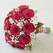 In this image, a classic romantic bridal bouquet of red roses and white gypsophila is captured in calm, diffused daylight, as if resting on a windowsill in a Tufnell Park townhouse. The roses are a deep, velvety crimson, their petals unfolding in neat spirals that draw the eye inward, each bloom full and confident yet tender in its openness. They are arranged in a rounded, hand-tied style, every flower facing slightly outward so the bouquet appears lush from every angle, much like a carefully composed centrepiece ready for an intimate reception near Fortess Road. Threads of gypsophila weave among the roses, the tiny white flowers forming a soft mist around the bolder blooms, giving the entire arrangement a feeling of lightness and breath. The contrast between the dense, plush roses and the airy, cloud-like sprays of baby's breath creates a tactile dialogue you can almost feel: strong, smooth petals set against the feathery touch of the filler flowers. The stems are gathered tightly and wrapped in an elegant ribbon, finished with a line of tiny pearls that catch the light in a subtle, sophisticated way, echoing bridal jewellery or a treasured accessory worn at a ceremony in nearby Dartmouth Park. Hints of greenery peek discreetly beneath the red and white, adding freshness without breaking the bouquet's romantic harmony. The overall mood is one of quiet celebration and enduring devotion, as though the bouquet has just been lifted from the hands of someone about to say I do in Tufnell Park.