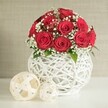 A dome of velvety red roses commands the centre of this image, creating a luxurious wedding table arrangement with a calm, romantic presence that feels at home in Tufnell Park's elegant terraces. The roses are tightly massed into a rounded bouquet, every bloom open and full, their petals layered in deep, wine-red spirals that seem to glow against the surrounding neutrals. Fine sprigs of white baby's breath are woven between the roses, their tiny blossoms forming delicate constellations that soften the bouquet and add an airy brightness. Here and there, small glimpses of fresh green foliage emerge, grounding the composition in nature without interrupting the dominant red-and-white palette. The flowers sit in a striking white vase, bulbous and rounded, constructed from intertwining strands that curl around one another in an open-weave pattern reminiscent of painted rattan or wicker. Light filters through the gaps in the weave, creating soft shadows on the smooth, pale tabletop beneath. To the left of the vase, two decorative orbs made from pale, woven material echo the same lattice effect, one larger sphere positioned slightly in front of a smaller one. Their natural beige tones bring warmth and texture, as if this table had been styled for an intimate celebration at a local venue just off Tufnell Park Road. Behind the arrangement, a gently blurred wall in light beige or cream serves as an unobtrusive backdrop, its subtle texture adding depth without stealing focus. The lighting is calm and even, suggesting daylight filtering through a nearby window, and it draws out every nuance of petal, weave, and shadow. The mood is one of understated luxury-ideal for wedding receptions, vow renewals, or romantic dinners set in and around Tufnell Park.