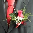A close-up photograph captures a romantic wedding boutonniere pinned to the lapel of a dark charcoal grey suit, styled as if for a ceremony in the heart of Tufnell Park. At its centre sits a partially unfurled rose bud in a rich, deep pink hue, somewhere between fuchsia and cerise, with petals that appear soft and velvety, gently spiralling towards the core. Surrounding the rose is a delicate ring of white gypsophila, each tiny blossom like a speck of cloud, adding a light, airy contrast to the bold central bloom. The florist has tucked in a combination of lush green foliage: broad, glossy leaves that lie smooth against the suit fabric, and slender, feathery sprigs that fan outwards, giving the arrangement a subtle sense of movement, as if it has just been gathered from a garden near Tufnell Park Fields. At the base, a bright red satin ribbon is tied into a neat bow, its reflective sheen catching the light in a way that echoes the plain red silk necktie beneath the jacket. The tie lies sharply aligned down the torso, framed by a grey dress shirt with a faint, elegant pattern that provides depth without distraction. The suit itself looks to be a fine wool blend, its tight weave appearing smooth and refined, a classic choice for grooms and wedding guests. The overall composition balances colour, texture, and form, creating a buttonhole that communicates care, celebration, and a polished sense of style suited to special occasions across Tufnell Park.