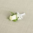 A classic white rose boutonniere is laid diagonally across a softly textured, light beige fabric that looks like natural linen, its fine crosshatch weave giving a calm, understated backdrop. At the heart of the piece sits a single creamy white rosebud, medium-sized and tightly furled, its velvety petals just beginning to open and catch the gentle, diffused light. Around the base of the bloom, hints of pale green sepals peek through, reinforcing the sense of freshness, as though the flower has just been prepared in a local Tufnell Park studio moments before heading to a nearby wedding on Fortess Road. Clusters of tiny white filler flowers, clearly reminiscent of baby's breath, nestle beside and just behind the rose, their cloud-like, starry blossoms adding an ethereal softness and a whisper of texture. Slender, pointed leaves in deep, vibrant green frame the rose and filler blooms, some smooth and glossy, others slightly more feathery, creating a natural cradle that balances the pure white tones. All the stems are neatly gathered and wrapped in a pristine white satin ribbon, the fabric pulled snug and even, then finished with a small, precise bow placed just below the flower head. The bow tails lie flat and orderly, suggesting a careful hand and practiced florist's skill, like the craftsmanship you'd expect when ordering wedding flowers for a ceremony near Tufnell Park Fields. The lighting is gentle and even, with no harsh shadows, so every petal, leaf, and thread of ribbon appears soft yet distinct, making this boutonniere feel both refined and quietly romantic-perfect for a groom or groomsman looking to add a subtle, traditional accent to a formal lapel on a North London wedding day.