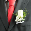 This close-up image reveals a classic white rose boutonniere resting against the rich charcoal fabric of an impeccably tailored suit, the kind you might see at a refined ceremony in Tufnell Park. The rose is perfectly formed and just beginning to unfurl, each velvety petal curling outward in a smooth spiral, glowing softly as though catching filtered daylight from a townhouse window near Yerbury Primary. Surrounding the rose, minute stars of baby's breath cluster tightly, their tiny white blossoms creating a cushion of softness that enhances the rose's purity without overwhelming it. Wisps of feathery greenery and looping, glossy leaves arc gracefully around the focal bloom, introducing lively shades of green that recall the tree-lined paths of nearby Hampstead Heath. At the stem's base, a narrow white satin ribbon is tied into a delicate bow, its subtle sheen and crisp lines speaking of careful, practiced hands and a deep respect for tradition. The boutonniere is positioned with precision on the suit's lapel, stitched in such a way that it lies flat yet feels full and dimensional, ready for a groom or prom guest to step into a day of photographs and speeches. The atmosphere it suggests is calm, polished, and emotionally charged, with a faint imagined fragrance of fresh rose petals and greenery that have just arrived from the market. Every detail, from the choice of rose variety to the angle of each fern frond, reflects thoughtful local craftsmanship, ideal for celebrations unfolding in and around Tufnell Park's churches, registry offices, and intimate reception venues.