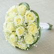 There is a serene, almost dawn-like hush to this bridal bouquet, assembled with the kind of craftsmanship I practise at Flowers Tufnell Park for couples marrying near Dartmouth Park or along Junction Road. From a soft overhead perspective the rounded dome of roughly fifteen to twenty creamy-yellow roses becomes the story's focus: each rose reveals a spiralled center and velvety outer petals that catch the light in subtle cream and ivory tones. Fine sprays of Gypsophila nestle in the gaps like morning mist, their tiny white stars giving the arrangement an ethereal, weightless quality. Curving blades of green - possibly lily grass - are woven around the edge, their sleek lines adding movement and a contemporary whisper to an otherwise classical silhouette. The stems are visible and neatly bound with pristine white satin ribbon, counterpointing the natural textures with a cool, elegant finish.

Resting on a pale, slightly textured linen surface, the bouquet benefits from soft, diffuse illumination that reveals micro-variations of cream and pale yellow in the petals and the gentle sheen on the ribbon. The scent is delicate and reassuring, the timeless rose perfume softened by green notes from the grasses and the clean air of an early wedding morning. This bouquet feels like quiet promise - designed for weddings in Tufnell Park, intimate ceremonies around Archway, or as an elegant anniversary gift - and is presented with the freshness and attention to detail local clients trust for same-day delivery in the neighbourhood.