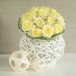 A refined floral centerpiece glows softly against a warm, neutral background, the kind you might find on a beautifully dressed wedding table in Tufnell Park. At its core is a rounded dome of light yellow roses, each bloom full and velvety, petals unfurling in layered whorls that range from creamy ivory at the edges to a mellow, buttery yellow at the centre. The roses are packed closely together, creating a sense of abundance and gentle luxury. Threaded between them, tiny white blossoms of baby's breath form airy clouds, their fine, branching stems introducing movement and delicate texture, like a whisper of springtime along Dartmouth Park Hill. A slender ring of fresh green foliage encircles the base of the flowers, neatly defining the bouquet and lending a subtle frame of colour. The arrangement is cradled in a sculptural white vase formed from interlaced strands that suggest wicker or rattan; its open, basket-like pattern allows light to pass through, casting lacy shadows onto the pristine white surface beneath. To the side, two understated decorative spheres, crafted from pale, natural-looking woven strips, echo the same openwork pattern and extend the visual rhythm established by the vase. The softly blurred beige backdrop remains unobtrusive, ensuring the focus stays firmly on the interplay of petals, weave, and light. The overall mood is one of modern elegance and calm joy, perfectly suited to wedding receptions, anniversary dinners, or stylish gatherings hosted in North London homes and local venues, with craftsmanship that feels intimately in tune with the area's relaxed sophistication.