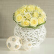 This arrangement is a soft study in ivory and pale yellow, a compact dome of creamy yellow roses hugged by dancing tendrils of gypsophila whose tiny white stars catch and scatter light. The bouquet rests atop a striking white wicker orb - a contemporary, tactile base whose airy latticework frames the flowers like a miniature pavilion. Petals are full and layered, their edges ruffled and slightly translucent when struck by afternoon sun, revealing warm inner tones that contrast gently with the cool white of the basket. The interplay of textures is deliberate: smooth rose petals, the feathery cloud of baby's breath, and the sculptural weave of the vessel invite both sight and touch. A faint fragrance drifts from the composition, a clean rose note tempered by a touch of earthiness, reminiscent of a Sunday walk through the local market or a picnic on the grassy slopes of nearby Dartmouth Park. Curved blades of greenery loop around the crown, adding a subtle, rhythmic punctuation that keeps the eye moving around the bouquet. In quieter interiors the piece reads as serene and hopeful - suited to a housewarming, a gentle congratulations, or the calming centrepiece at an intimate celebration on a balmy evening. As a local florist familiar with the light and pace of Tufnell Park, I arrange each stem to balance brightness and restraint so the bouquet feels both modern and tender, a small sculpture of comfort and understated joy.