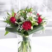 A luxurious red and white bouquet stands proudly in a clear cylindrical glass vase, its neatly trimmed green stems visible through the fresh water, suggesting the care and precision of an experienced Tufnell Park florist. Deep red roses sit at the heart of the design, their velvety petals just beginning to unfurl with a sense of classic romance and quiet drama. A large, ruffled crimson carnation nestles at the front, its tightly layered petals creating a rich, tactile focal point. Around these, creamy white alstroemeria with fine speckles, white chrysanthemums, and closed white lily buds offer a softer counterpoint, promising future blooms and a lingering sense of anticipation. Wisps of delicate baby's breath float between the larger flowers like tiny stars, adding lightness to the composition and softening the bold reds. The bouquet is cradled by an abundance of lush greenery: broad, glossy dark green leaves are wrapped and gently curved around the base, forming an elegant collar, while slimmer, eucalyptus-like stems and pointed foliage stretch upward to add height and texture. The arrangement rests on a bright white surface, set against a softly blurred, light background that could easily be a sunlit Tufnell Park townhouse window or a calm corner of a local office. Natural, even daylight bathes every petal and leaf, enhancing the vivid crimson tones and clean whites, and giving the bouquet an air of celebration and thoughtful intention. The overall mood is sophisticated yet welcoming, ideal for marking an anniversary, a special birthday, Mother's Day, or simply sending warm congratulations to someone in and around Tufnell Park.