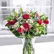 A luxuriant bouquet of deep red roses, matching red carnations, white alstroemeria, and clouds of tiny white gypsophila stands proudly in a clear glass vase on a spotless white table, bathed in soft daylight from a large window behind. Each crimson rose, some fully open with velvety petals spiralling outward and others still in elegant bud, forms the dramatic heart of the arrangement, while ruffled red carnations nestle between them, adding richness and volume. Clusters of white alstroemeria, many still in tight buds with just a hint of green at the tips, are interspersed throughout, their partially open blooms revealing delicate, trumpet-shaped petals that lighten the composition. Sprays of baby's breath create a fine, mist-like layer, softening the bold reds with a gentle, airy texture that looks almost like a dusting of snow. A generous collar of broad, glossy dark green leaves wraps around the base, while slimmer, lighter green foliage threads upward through the flowers, giving movement and depth. Through the sides of the vase, the neatly cut stems can be seen resting in clear water, emphasising the bouquet's freshness, as if it has just arrived from a local florist in Tufnell Park. The background outside the window fades into a pale, almost monochrome urban scene, reminiscent of a quiet street off Fortess Road, deliberately blurred so that the vibrant reds, whites, and greens appear to glow. The overall effect is one of joyful elegance, ideal for marking a romantic evening, a special birthday, or simply bringing warmth into a North London home on a grey day.