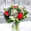 A contemporary mixed bouquet glows in a clear glass vase, captured in soft daylight as though set beside a bright window in a Tufnell Park townhouse. At the heart of the arrangement, a coral-red rose and a deeply ruffled crimson carnation draw the eye, their velvety petals layered and plush, radiating a feeling of quiet passion and warmth. Clusters of delicate white chrysanthemums and tiny daisy-like blooms gather around them, each petal crisp and neat, adding a gentle, cloud-soft contrast against the bold reds. Slender stems of creamy white lilies, some still in tight bud, rise gracefully from the centre, their pale green tips suggesting blossoms that will slowly open over the next few days, extending the life and changing beauty of the display. Around the base, broad, glossy leaves in deep emerald green are artfully folded and wrapped in sweeping arcs, cradling the flowers and giving the bouquet a sculpted, modern silhouette that feels both elegant and welcoming. The glass vase is simple and clear, revealing straight, carefully trimmed stems and clean water, suggesting freshness and expert care, much like arrangements prepared daily in local studios near Tufnell Park station and along Fortess Road. Natural light washes over each bloom, picking up subtle shadows and highlights, as if the bouquet is resting on a calm dining table ready to greet guests. The mood is uplifting yet composed, ideal for celebrating a special birthday, saying thank you to a neighbour on Carleton Road, or bringing a touch of brightness and texture to an otherwise ordinary afternoon at home.
