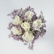 A high-angle overhead photograph shows a compact, hand-tied bouquet arranged in a perfect circle, laid on a smooth, pale grey surface that could easily be a kitchen table in a Tufnell Park townhouse. At the very centre, six creamy white roses dominate the view, each bloom tightly cupped yet beginning to unfurl in soft spirals. Their petals look velvety and cool to the touch, with a subtle ivory sheen that catches the light, while the outer edges remain crisp and clean. At the base of some petals and sepals, the faintest wash of pale green adds a natural freshness, like new growth after rain on Dartmouth Park Hill. Surrounding the roses is a dense collar of lilac and muted lavender limonium, sometimes called statice, forming a soft halo of tiny clustered flowers. These miniature blossoms create a feathery, cloud-like texture that contrasts beautifully with the smooth, substantial rose petals, giving the bouquet an almost weightless feel. The filler flowers are tightly packed, providing a purple mist that makes the white roses glow even more vividly, as if they are floating above a field of heather. The lighting is even and gentle, without strong shadows, so every curve and fold of the petals is clearly defined yet soothing on the eye. This bouquet feels calm, thoughtful, and refined, perfectly suited for sympathy flowers, a quiet anniversary, or a comforting delivery to a friend off Fortess Road. The overall mood is serene and romantic, as though the flowers are pausing in a moment of stillness before being placed tenderly into someone's hands.