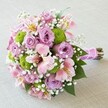 A vibrant, lovingly composed bridal bouquet glows atop a gently textured, pale surface, recalling the tranquil ambiance of Tufnell Park's hidden gardens. Its compact, rounded design features a harmonious layering of lush lavender and blush-pink roses, each bloom opening in soft whorls that radiate charm and understated elegance. Dotted throughout are perky lime green button mums, reminiscent of fresh spring buds emerging near the wide avenues of Tufnell Park. Delicate Alstroemeria lilies, their trumpet-shaped petals kissed with pink, yellow, and burgundy, add a graceful, airy dimension, their individuality mirroring the local community's character. Ethereal white clouds of baby's breath spiral through the arrangement, softening the palette with a gentle mist-like quality. Forest-green foliage peeks from beneath the blossoms, bringing fullness and vibrancy reflective of the area's leafy parks. The bouquet's stems are tightly bound and wrapped with a shimmering fuchsia ribbon, thoughtfully adorned with a line of small pearl beads-evoking celebrations and heartfelt commitments. This sophisticated yet joyful bouquet would delight any bride, light up an anniversary celebration, or mark an engagement with contemporary flair in Tufnell Park. Its colour story and delicate texture highlight the fresh, local craftsmanship, embodying the romance and beauty that define floral artistry in this North London neighbourhood.