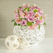 This image captures an elegant, rounded floral arrangement styled as though it's about to grace the table of a small wedding breakfast in Tufnell Park. The bouquet is tightly composed, forming a neat dome where pastel tones melt gently into one another. Lavender and rich pink roses sit at the core of the design, their open blooms layered with velvety petals that appear almost luminous under the soft, even light. Between them, slender stems of pale pink alstroemeria weave in and out, their trumpet-shaped blossoms etched with fine speckles that echo the delicate markings you might notice on wildflowers in nearby Tufnell Park playing fields. Clusters of lime green button chrysanthemums offer a vivid burst of colour and a pleasingly tactile texture, each button head adding rhythm and a contemporary flair that prevents the pastels from becoming too sweet. Tiny sprays of white baby's breath drift through the spaces between the larger flowers, creating a gentle haze and a sense of lightness, while discreet dark green foliage provides a calm, natural frame. The entire composition rises from a round white vase made from intricately woven wicker or rattan, its open, basket-like lattice forming a sculptural globe that feels both modern and organic. Resting on a pristine white tabletop, the vase is flanked by two smaller woven spheres in a lighter, natural tone, their broad, flat strips arranged in loose open patterns that echo the main vessel. A softly textured, neutral wall behind ensures nothing distracts from the bouquet. The overall mood is tranquil and refined, perfectly suited to wedding décor, registry office celebrations, or romantic dinners at home in the Tufnell Park area, with the flowers looking fresh enough to have just been arranged that morning.