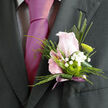 The image captures a romantic boutonnière designed with the care of an experienced Tufnell Park wedding florist, laid neatly against a deep, tailored lapel. At the centre sits a blush-pink rose, not fully open, its layered petals cupping inward as if holding a secret, their colour somewhere between ballerina slipper and the first streak of dawn over Tufnell Park Fields. Encircling this focal bloom, sprigs of white gypsophila extend in soft, cloud-like clusters, each minuscule flower adding a sense of airiness and gentle movement, like breath on a cool morning. Tucked alongside the rose, a single green button chrysanthemum adds a sharp, contemporary note, its rounded form and vibrant lime hue giving a refreshing contrast to the muted pinks and whites. Fine strands of greenery and a narrow blade of sleek foliage arc behind the flowers, shaping the silhouette so the arrangement sits cleanly on a suit jacket without feeling bulky. The stems are bound in a pale mauve ribbon, satin-like and slightly reflective, wrapped in tight, even coils that hint at the florist's practiced hands and attention to detail. You can almost imagine the soft rustle as the ribbon is tied and the quiet satisfaction when the pin is set just right, ready to be worn to a wedding at a nearby town hall or a celebration in a tucked-away restaurant off Junction Road. The overall mood is tender and understated, perfect for prom nights, civil ceremonies, or intimate gatherings across Tufnell Park, where a single rose worn at the chest can say more than any number of words.