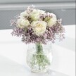 A close-up photograph shows a tranquil, beautifully balanced bouquet featuring creamy white roses and clouds of soft lavender-purple statice, captured in gentle daylight as if on a calm morning in Tufnell Park. The roses sit proudly at the heart of the arrangement, their petals unfolding in smooth, velvety spirals, with the faintest blush of pale yellow nested at the centre of each bloom, giving a sense of freshness and quiet luxury. Around them, the statice forms a misty halo of tiny, papery flowers, their lilac tones adding both colour and texture, like a lavender haze drifting across the frame. Slender green stems and a few subtle leaves peek through the mass of flowers, grounding the design with natural structure. The bouquet rests in a clear glass vase with a distinctive crackled, ice-like surface; the texture catches and scatters the light, sending tiny reflections across the bright white tabletop beneath. Through the water-filled vase, the green stems are clearly visible, straight and clean, suggesting flowers that have just been prepared by a careful florist. The background fades into a soft grey blur, with no distractions, so all attention remains on the blooms and the shimmering glass. The atmosphere of the image is peaceful and contemplative, like a quiet moment looking out toward Hampstead Heath from a Tufnell Park townhouse window. It feels perfectly suited for a thoughtful get-well gift, a gentle condolence gesture, or simply bringing a sense of calm, understated elegance into a home office or reading nook near Fortess Road, where subtle beauty and serenity are appreciated.