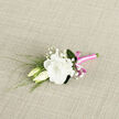A delicate boutonniere lies horizontally on a softly textured, light-coloured fabric, as if waiting to be pinned before a ceremony in one of Tufnell Park's characterful venues near Tufnell Park Road. The central flower is a lush white lisianthus, its petals velvety and gently ruffled, forming a cup of creamy softness that suggests both purity and warmth. Close beside it, two lisianthus buds lean inward, one tight and streamlined, the other just beginning to open, like a quiet promise of what is yet to come. Sprigs of baby's breath weave between the blooms, each tiny white floret contributing a fine, cloudlike texture, softening the transitions between flower and foliage. Slender, reed-like green leaves and tendrils extend from behind the main bloom, arching slightly and adding a sense of movement, as though a breeze from nearby Hampstead Heath had just brushed past. The stems of the flowers are carefully gathered and wrapped in a vivid magenta-pink satin ribbon, its surface smooth and reflective, catching the light with a refined sheen. A neat, compact bow rests at the front, introducing a cheerful, modern accent that contrasts beautifully with the understated ivory and green palette. The background fabric appears woven and natural, its subtle pattern enhancing the organic elegance of the arrangement without drawing focus away from the florals. This boutonniere feels ideal for a Tufnell Park wedding, civil partnership, or prom, offering a small but expressive flourish that speaks of attention to detail, gentle romance, and the craft of an experienced local florist who understands how flowers complement both person and place.
