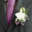 A sharply focused close-up of a groom's charcoal grey suit in Tufnell Park, showing a meticulously tailored jacket, patterned dress shirt, and a vibrant magenta silk tie framed by crisp lapels. Pinned to the left lapel is an elegant white lisianthus boutonnière, its fully opened bloom ruffled and soft, like layers of fine tissue, edged with a gentle cream tint. Two slimmer ivory buds lean protectively over the main flower, captured just before they unfurl, while airy clouds of white gypsophila scatter around them, their tiny blossoms catching the light like droplets of morning mist over nearby Hampstead Heath. Fine, linear greenery stretches outward in graceful arcs, giving the buttonhole an almost weightless, dewy freshness against the deep matte of the wool suit. At the base, a broad, glossy green leaf is wrapped snugly around the stems, secured with a narrow mauve ribbon that echoes the rich colour of the tie and adds a whisper of romance without overwhelming the clean, modern look. The lighting is soft and even, highlighting each petal vein, each thread in the fabric, and the careful craftsmanship that went into wiring and binding the flowers. The image feels poised on the brink of a significant moment-perhaps a wedding ceremony at a townhouse venue off Fortess Road or an intimate celebration near Tufnell Park station-capturing the sense of quiet anticipation, new beginnings, and heartfelt promise carried in this single, beautifully composed floral detail on the lapel.