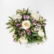 A top-down view of an elegant hand-tied bouquet shows creamy white and soft ivory roses gathered closely together, each bloom at a different stage of opening so that tight buds, half-open spirals, and fully unfurled petals create a gentle rhythm across a brilliant white background. Nestled between the roses are stems of alstroemeria, their slender buds and opening flowers painted in mauve, lavender, and purplish-pink, some petals etched with fine green streaks that add a delicate, painterly detail. Tiny white clouds of gypsophila float between the larger flowers, their airy clusters softening every edge and giving the whole arrangement a light, romantic haze. A collar of deep, glossy green foliage frames the bouquet, broad leaves curving protectively around the blooms and emphasizing their freshness and softness through contrast. The lighting is bright and clean, echoing the calm, contemporary interiors you might find in a townhouse just off Tufnell Park Road, and there are no visible ribbons or vases to distract from the flowers themselves. The overall impression is one of balance and quiet luxury, as though the bouquet has just been set down on a white table in a sunlit flat near Tufnell Park station, ready to mark an anniversary, a thoughtful birthday, or a peaceful moment of sympathy. Every detail suggests the skilled hands of a local florist who understands how London light plays on petals, crafting something both refined and heartfelt for delivery across Tufnell Park and the neighbouring streets.